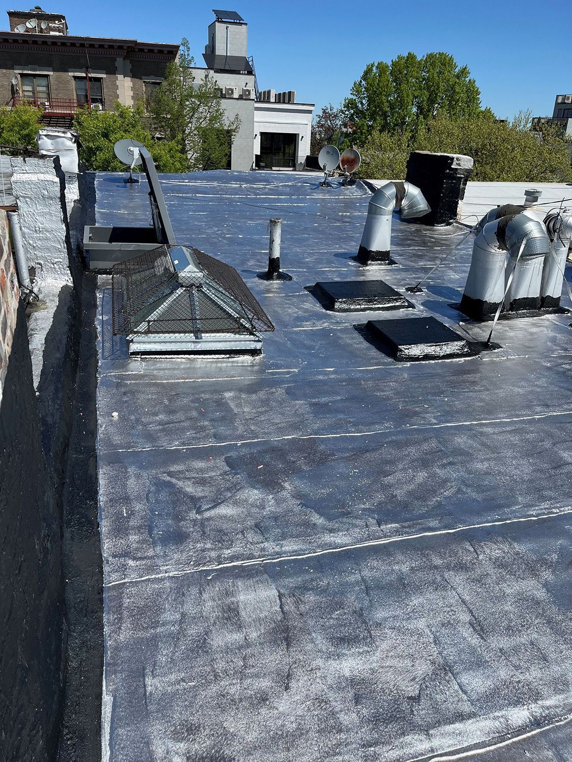 Rooftop view of a dark, flat tar roof with various vents, a skylight, and antennas under a sunny sky.