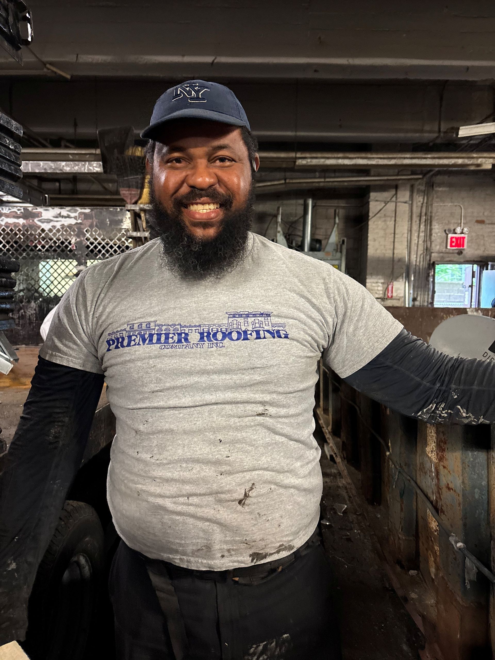 Smiling man in a gray shirt and cap, standing indoors near industrial equipment.