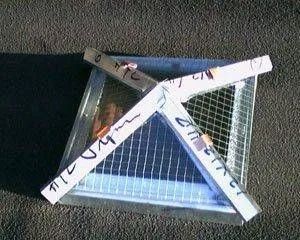 A square wire mesh covered window with white taped cross over it, sitting on pavement.