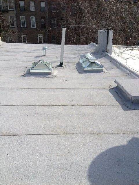 Flat rooftop with two skylights, vents, and a tall building in the background.