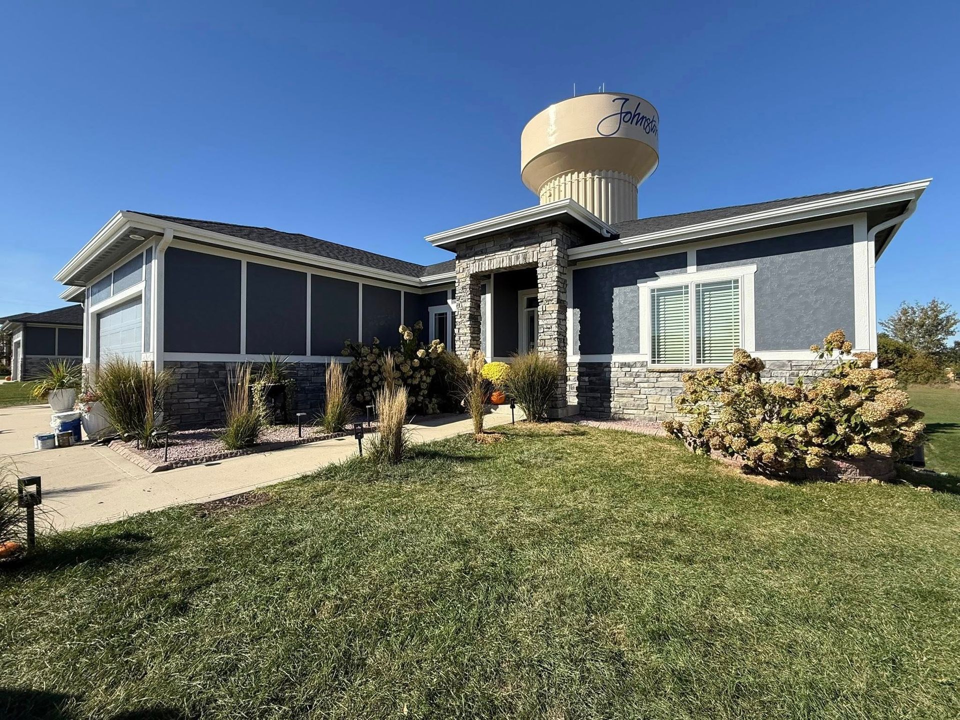 A blue single-story house with stone trim and a visible water tower behind the entrance, under a clear blue sky.