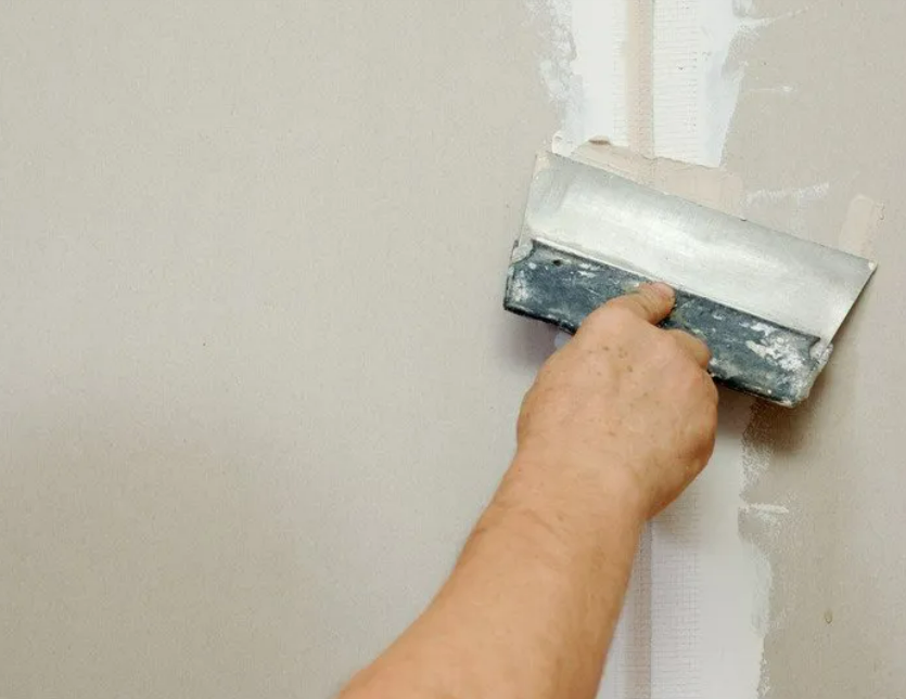 Hand using a trowel to apply drywall compound to a wall joint, beige wall.
