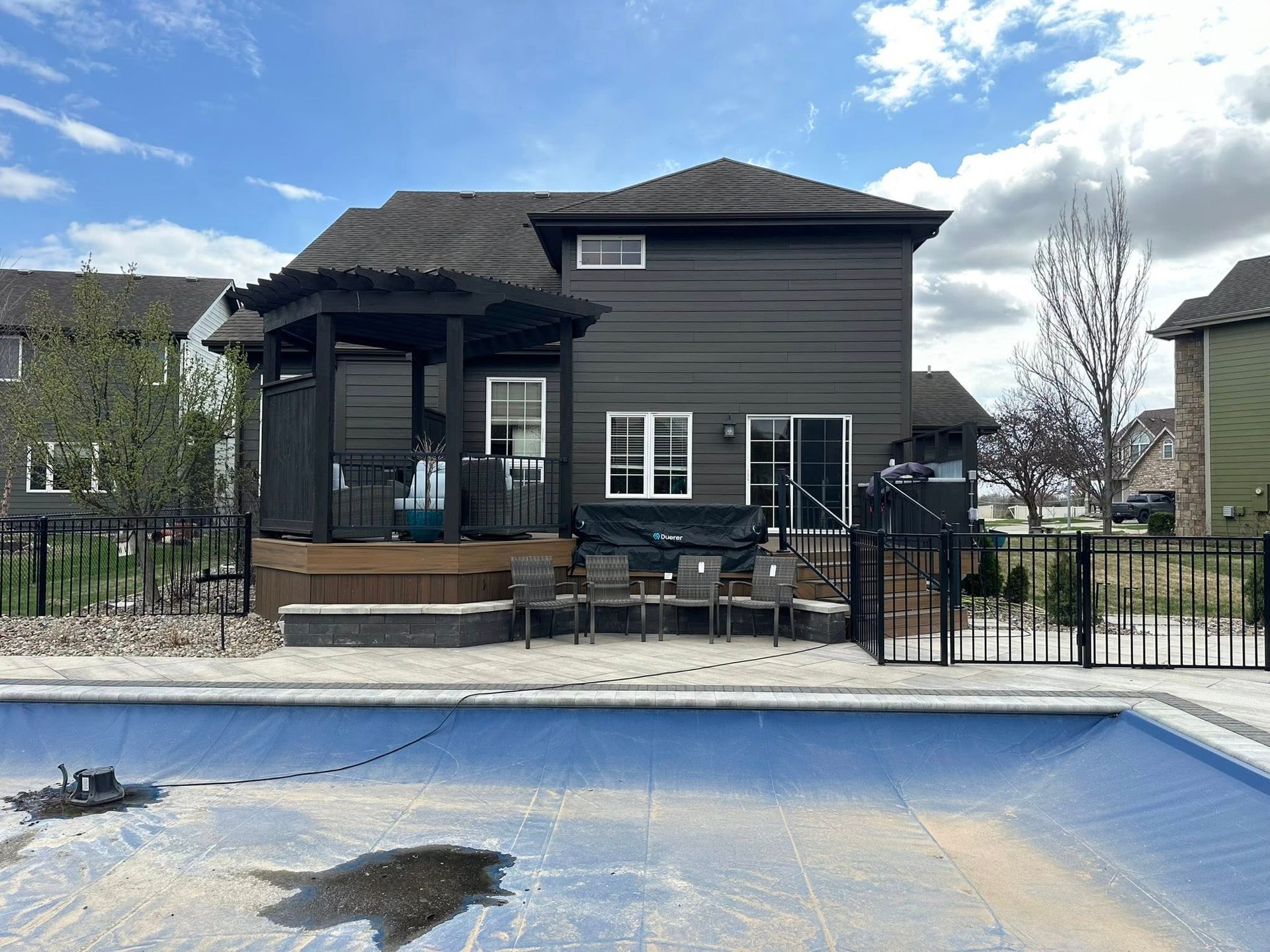 Dark house with pool, black fence, and deck; chairs sit near pool with partially open cover.