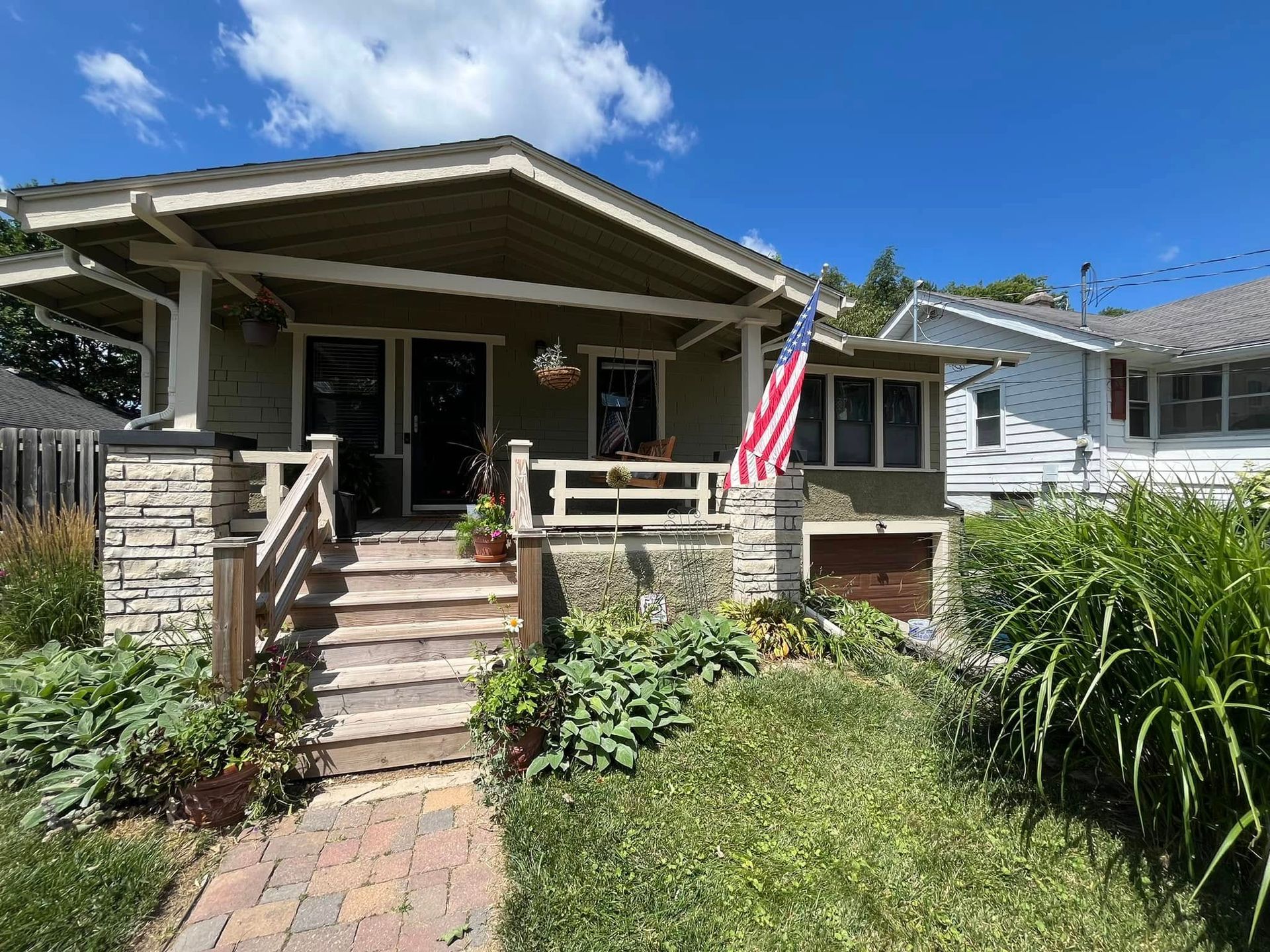 Bungalow-style home with stone accents, covered porch, American flag, and landscaping under a blue sky.