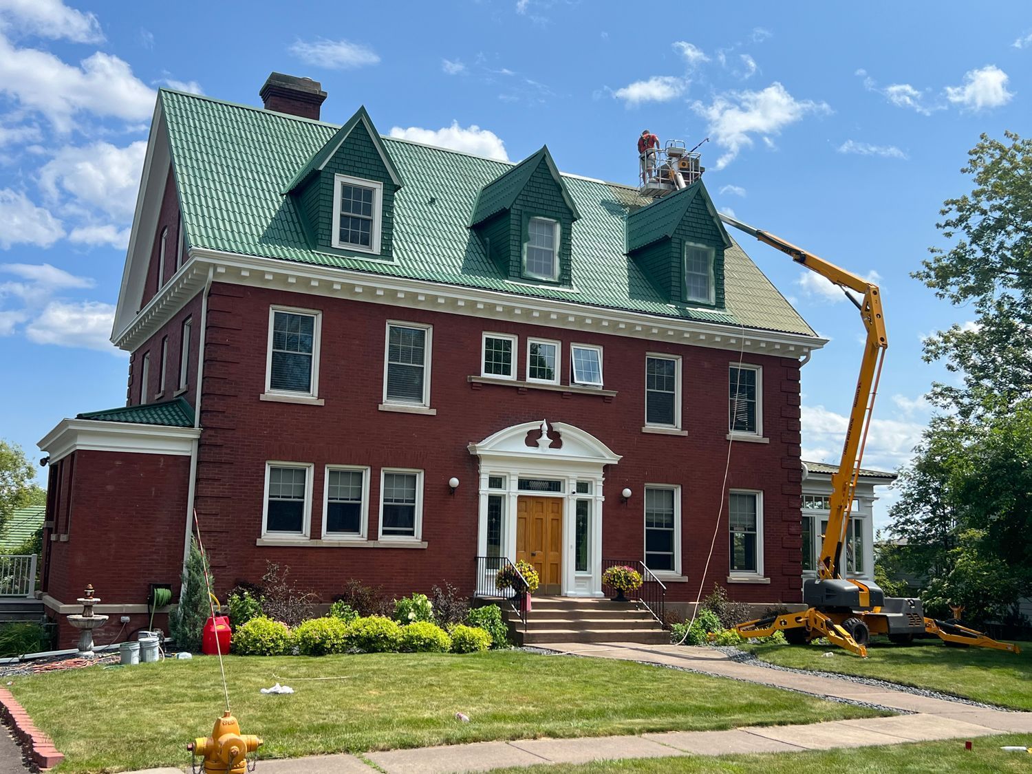 Red brick house with green roof; workers on roof with a yellow lift.