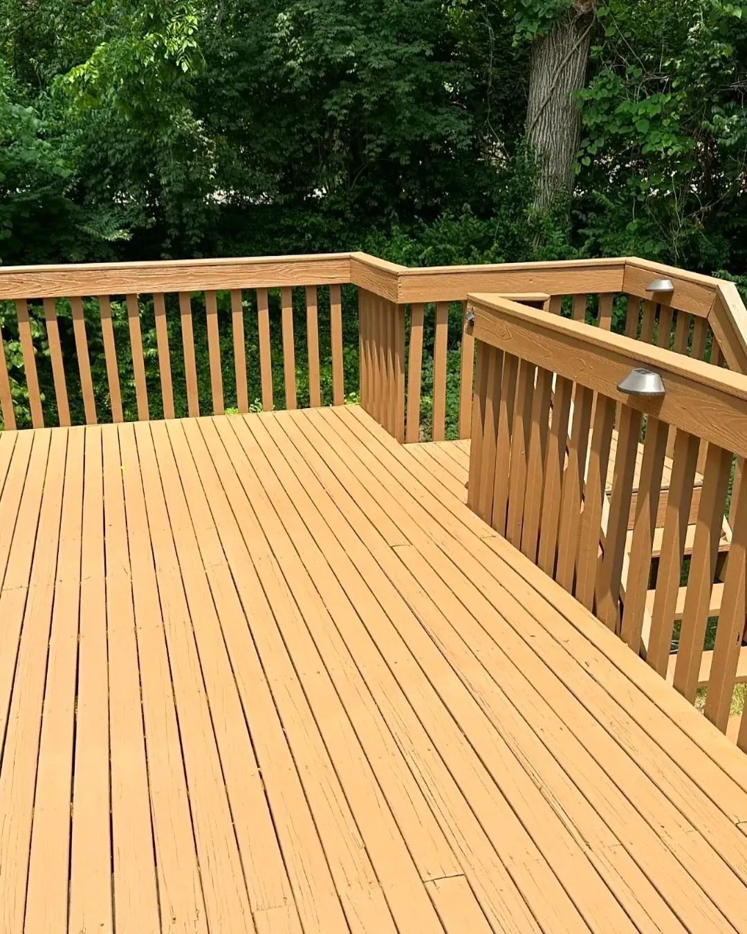 Wooden deck with railings, painted tan, surrounded by lush green trees.