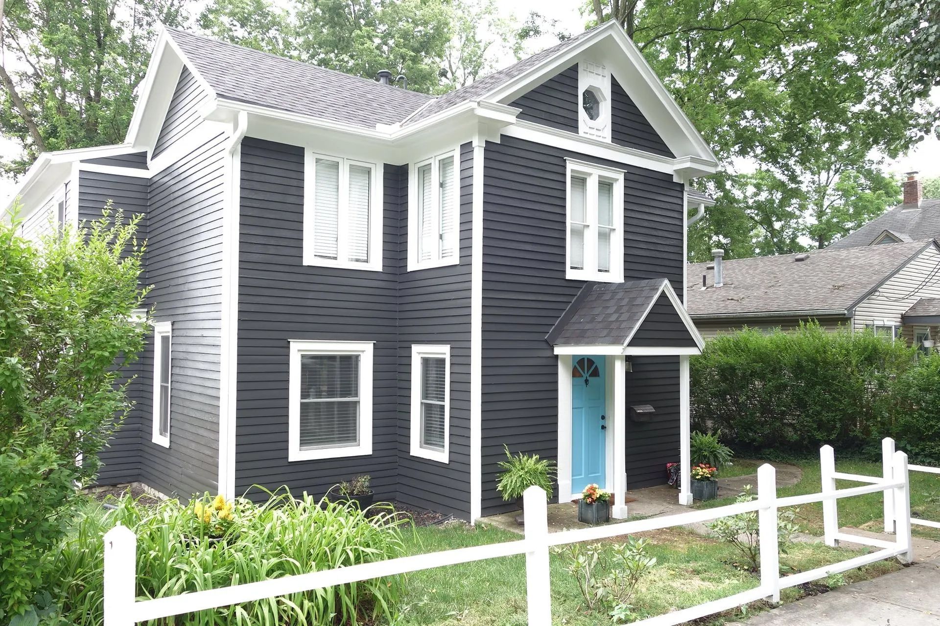 Black two-story house with white trim and a blue door, behind a white picket fence.