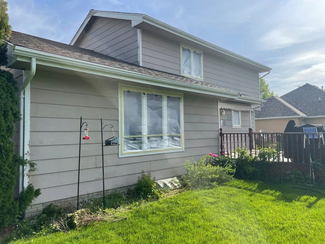 Exterior view of a two-story house being painted, tan siding, white trim, windows covered with plastic, green grass.