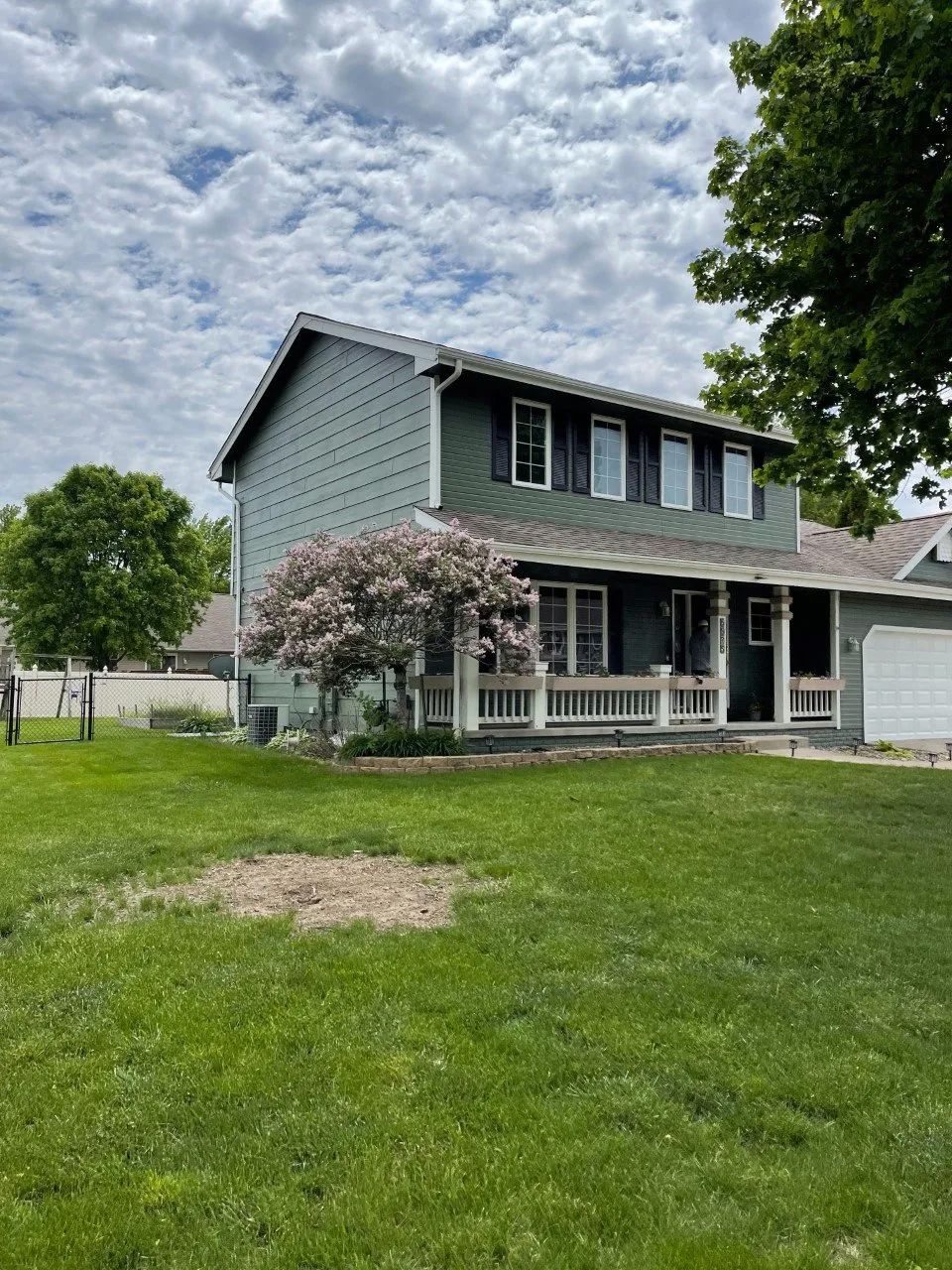 Two-story green house with a porch and a flowering tree in front. Blue sky with clouds.