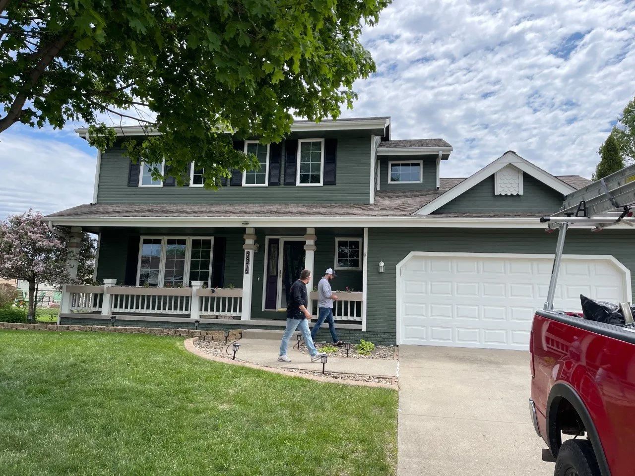Two-story green house with people in front; red truck with ladder parked next to the garage.