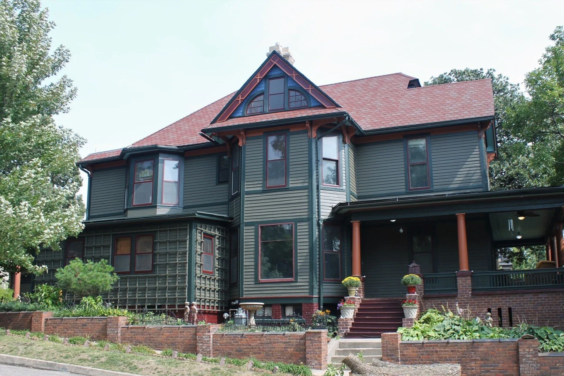 Green Victorian house with red roof and trim, set in a brick-lined yard, trees on either side.