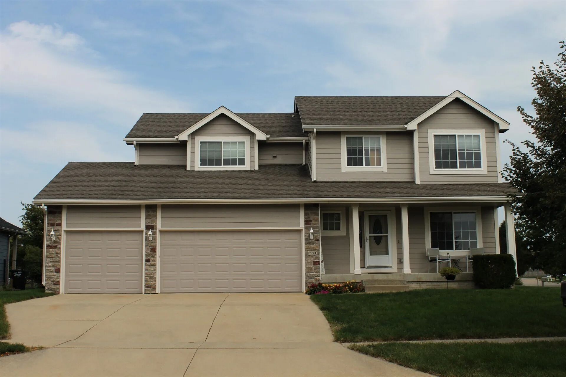 Two-story house with gray siding, three-car garage, and covered porch under a cloudy sky.