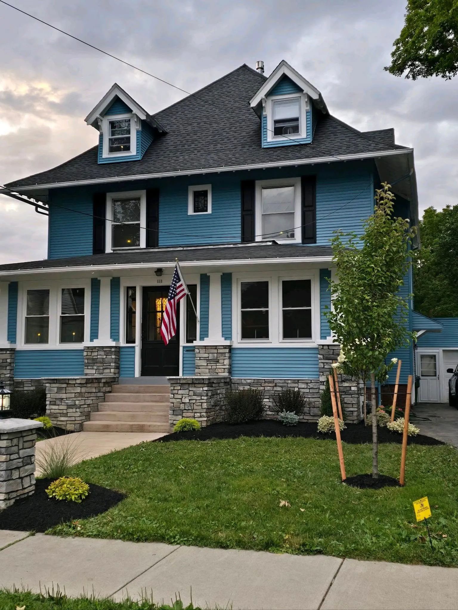 Blue house with black shutters, American flag, stone accents, and dormers.