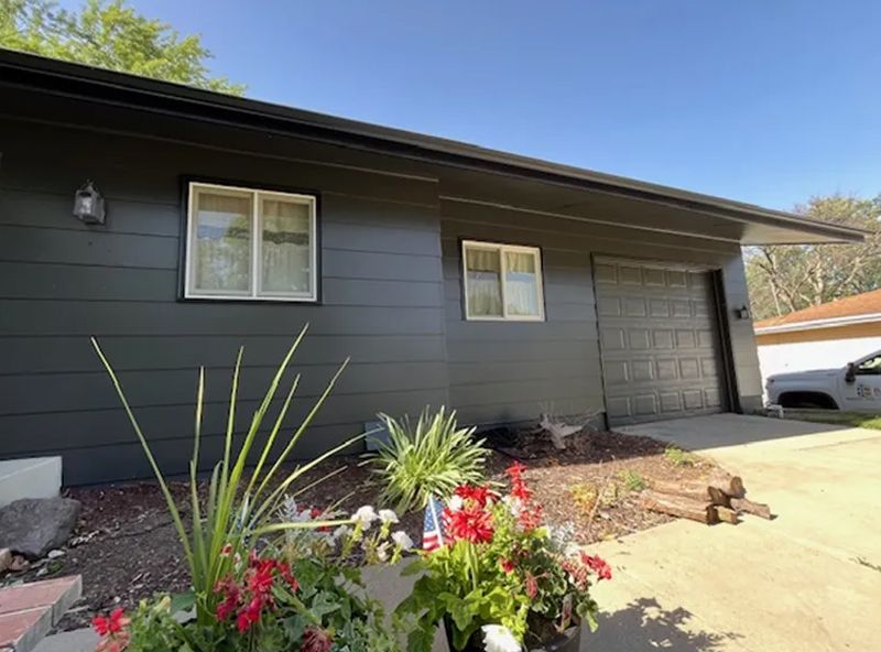 Black-painted house with white-framed windows and garage door, flower bed, and a bright sunny day.