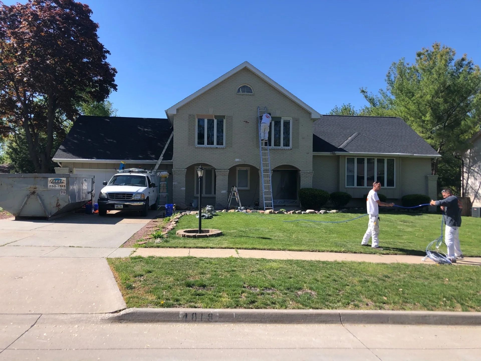 House exterior being painted by three workers; one on ladder, two on lawn. Sunny day.