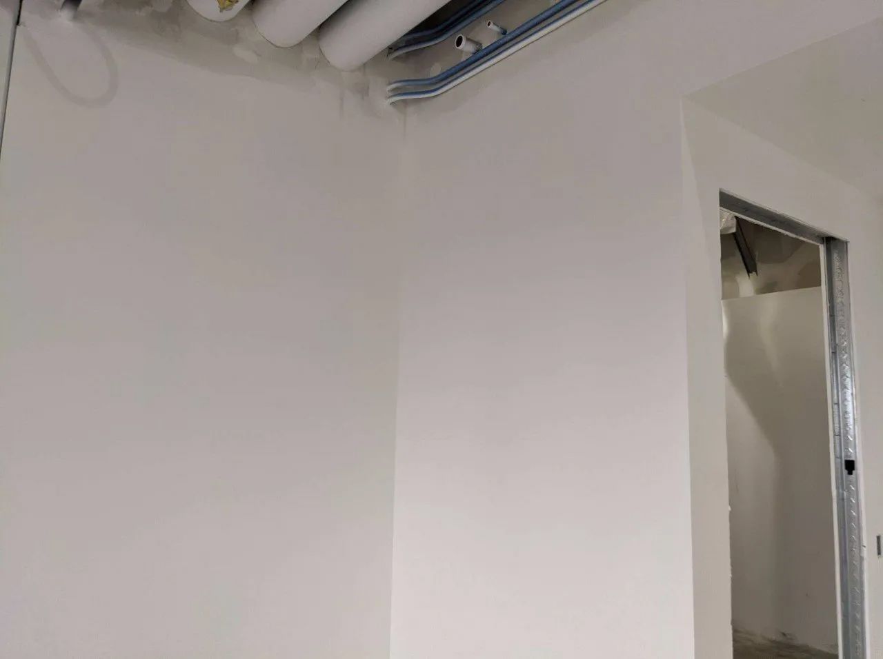 White walls in a room under construction, with exposed pipes and a doorway with metal framing.