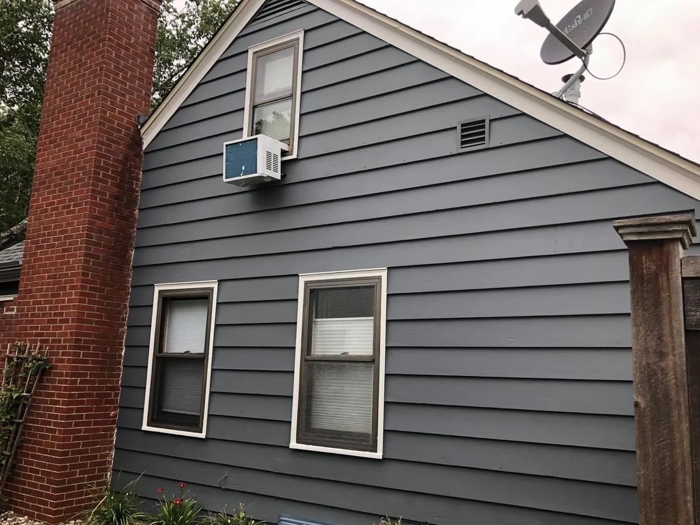 Dark blue siding on a house with an air conditioner in a window, chimney on the left, and satellite dish on the roof.