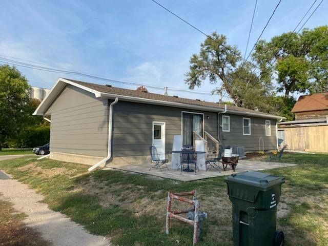 Back of a gray house with a small patio, green yard, and a trash can in the foreground.