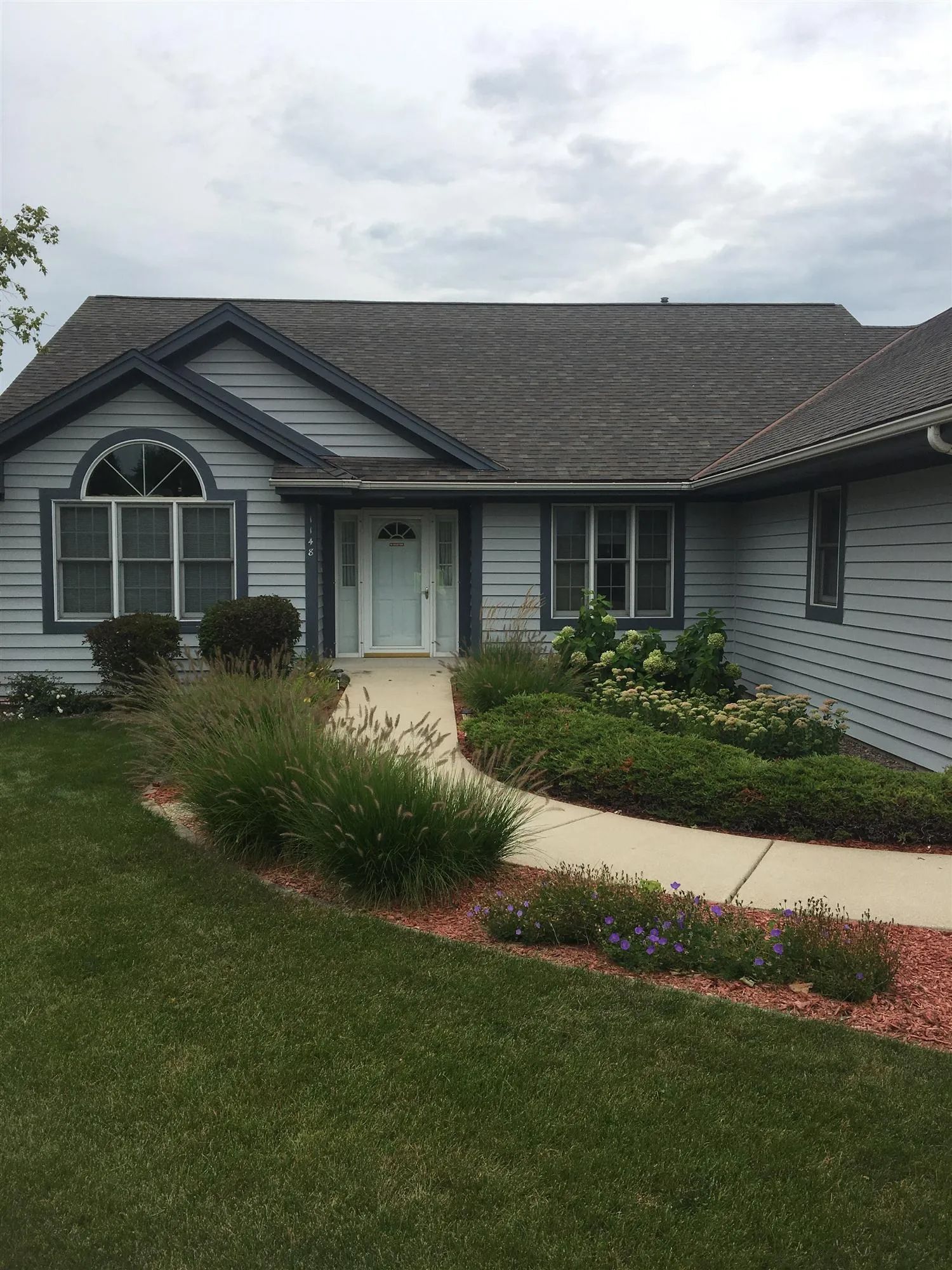 A light blue house with a curved path leading to a white door, surrounded by landscaping under a cloudy sky.