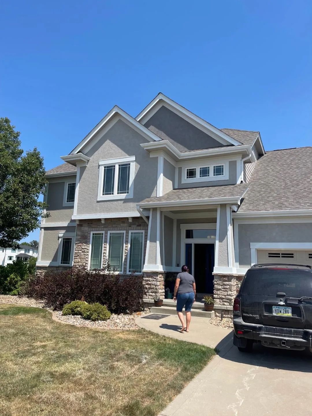 Woman walks towards a two-story house with stone accents and a gray exterior under a blue sky.