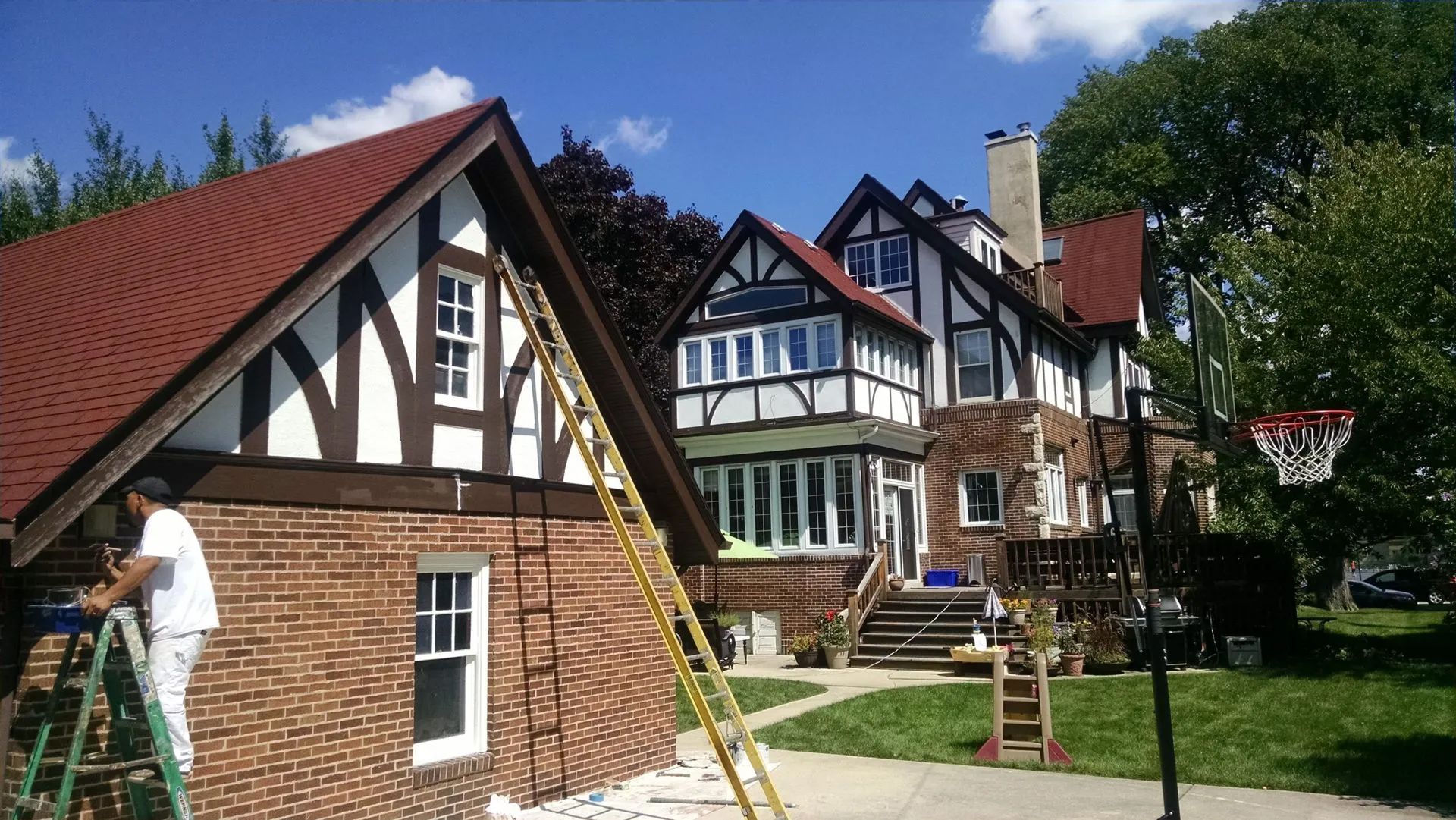 Man painting brick wall of a Tudor style house, with brown and white trim. Basketball hoop in yard.