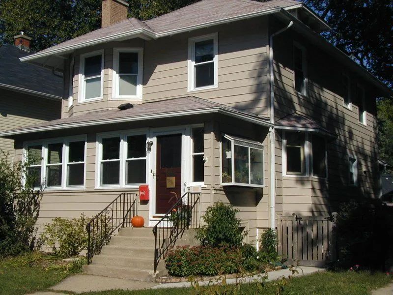 Two-story tan house with porch, steps, dark door, and small landscaping.
