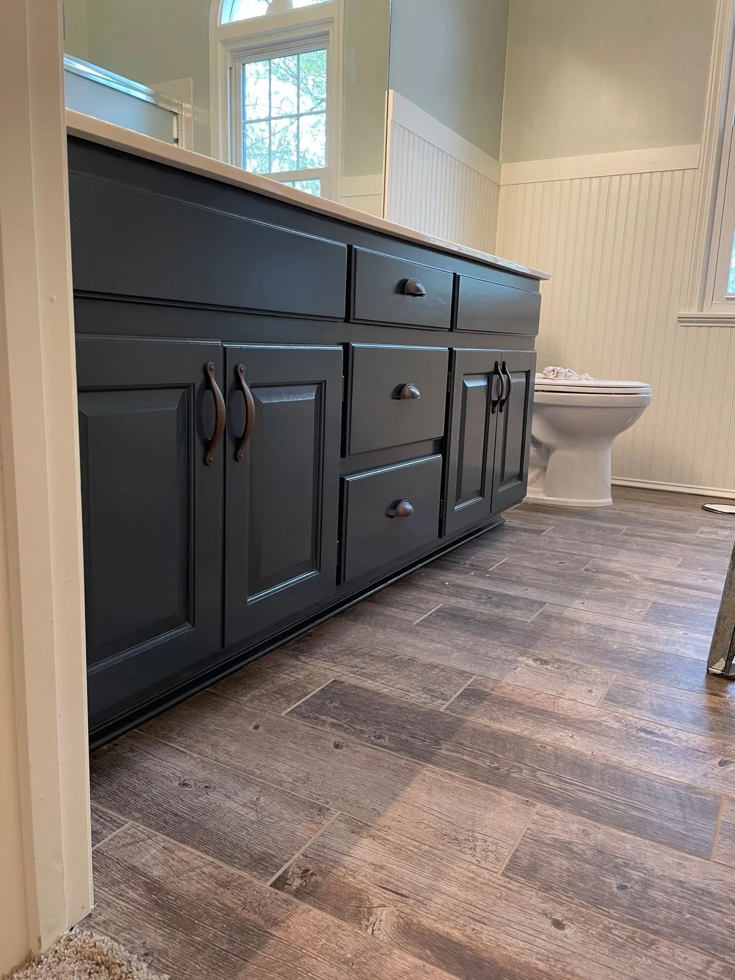 Dark blue bathroom vanity with bronze hardware, gray and brown wood-look flooring, white toilet.