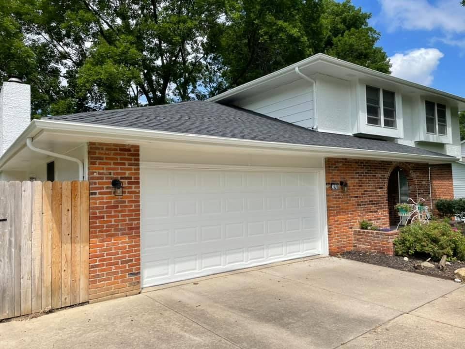 Two-story house with white garage door, red brick accents, new black roof, and green trees.