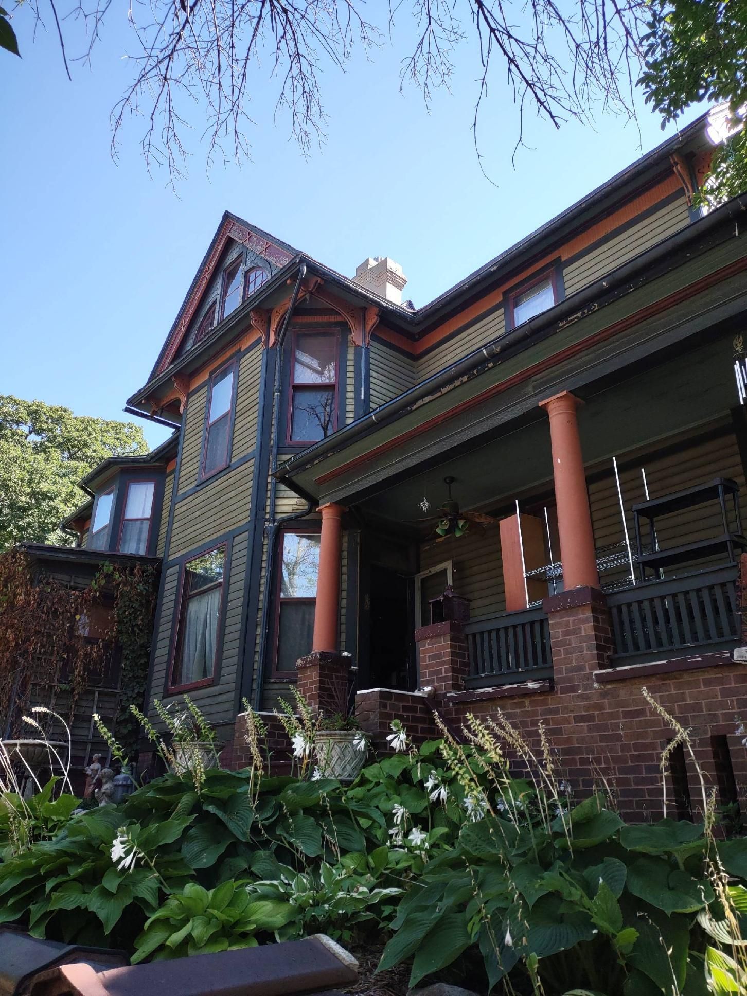 Victorian house with green siding, orange trim, and a porch, viewed from a garden on a sunny day.