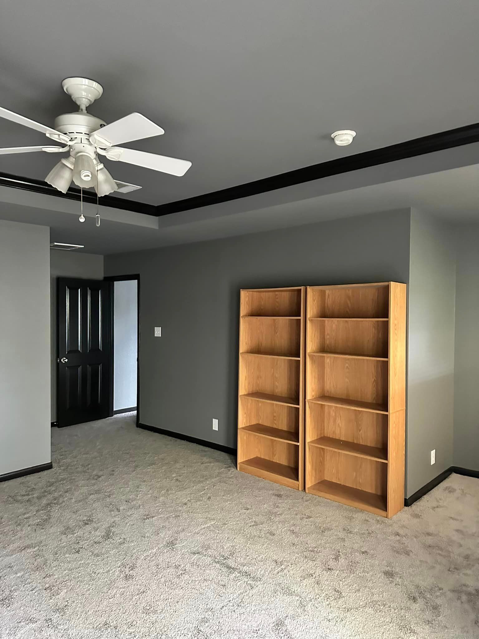 Bedroom with gray walls, ceiling fan, black trim, two oak bookshelves, and a dark door.