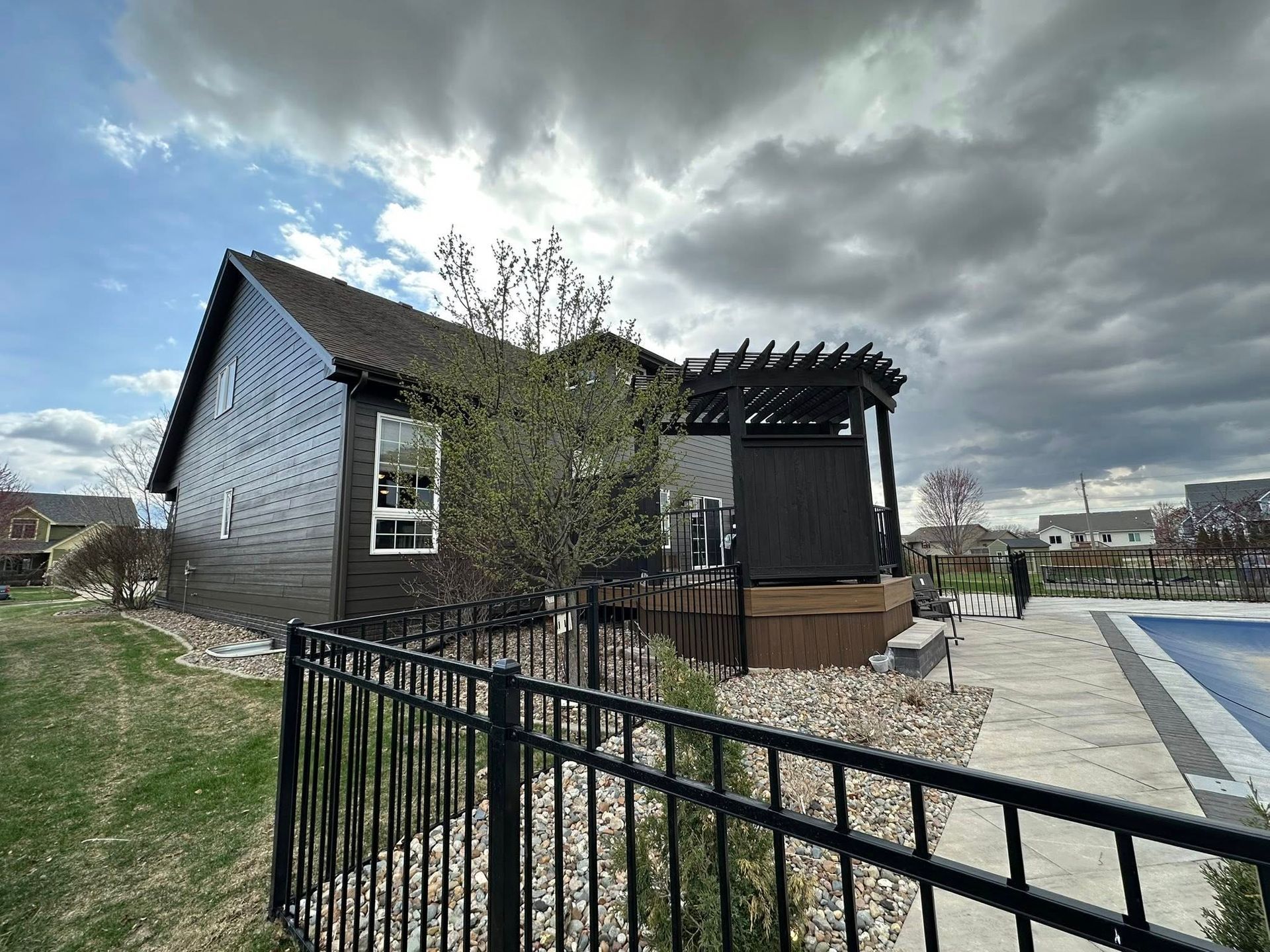 Black house with a deck, pergola, and fence, near a pool under a cloudy sky.
