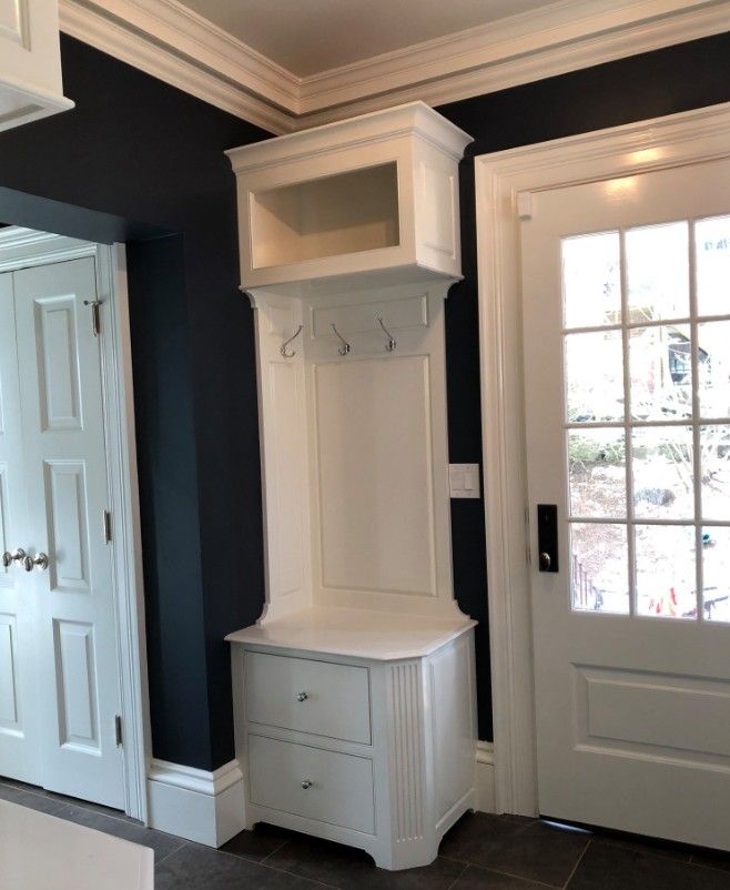White built-in entryway storage with hooks, drawers, and cabinet, against dark blue wall, near a white door.