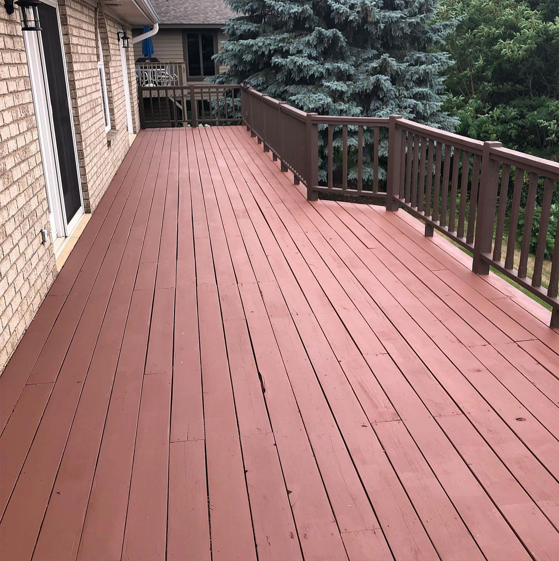 Brown wooden deck attached to a brick building, surrounded by a brown railing, and lush green trees.