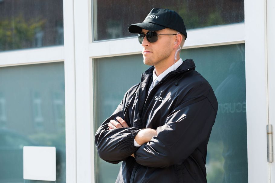 A security guard, wearing sunglasses and a black jacket, stands with arms crossed in front of a glass door.