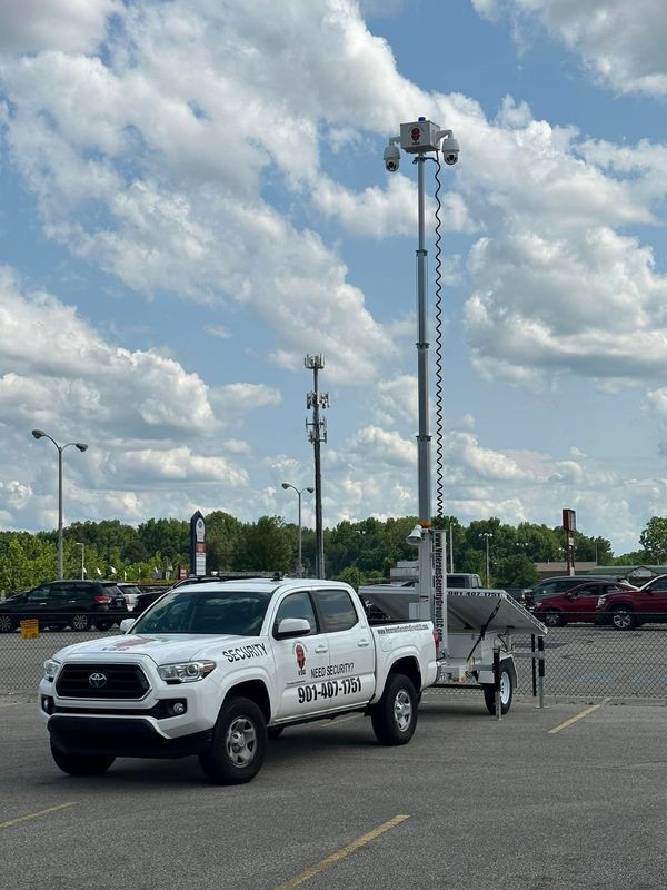 A white Toyota truck towing a trailer with a tall security camera tower in a parking lot on a partly cloudy day.