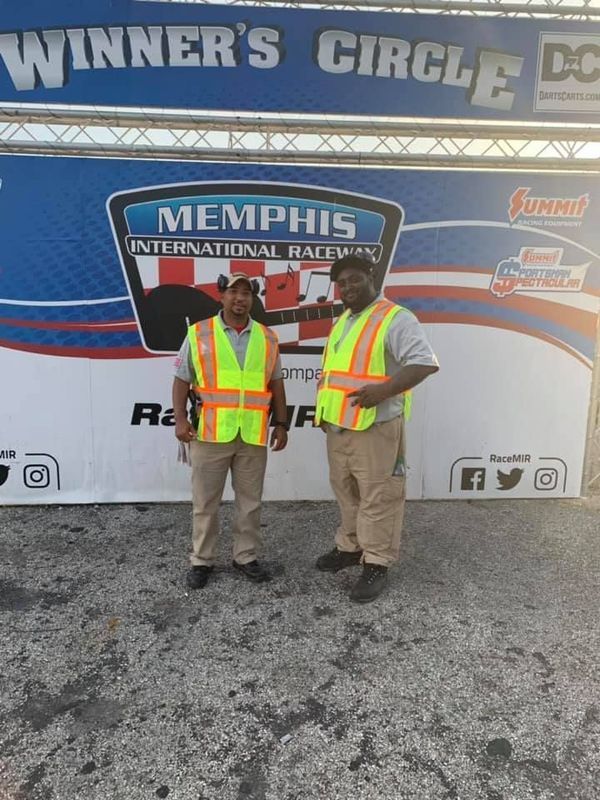 Two men in safety vests stand in front of the Memphis International Raceway Winner's Circle sign.