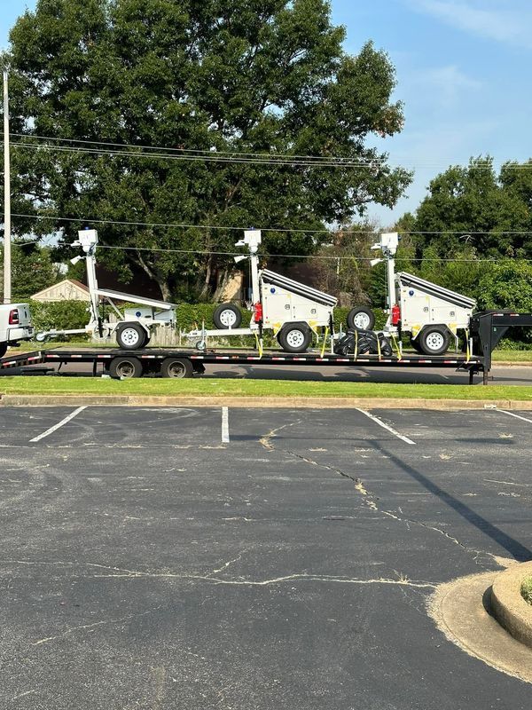 A trailer carrying three white mobile solar light towers parked in a parking lot. Trees and a clear sky are in the background.