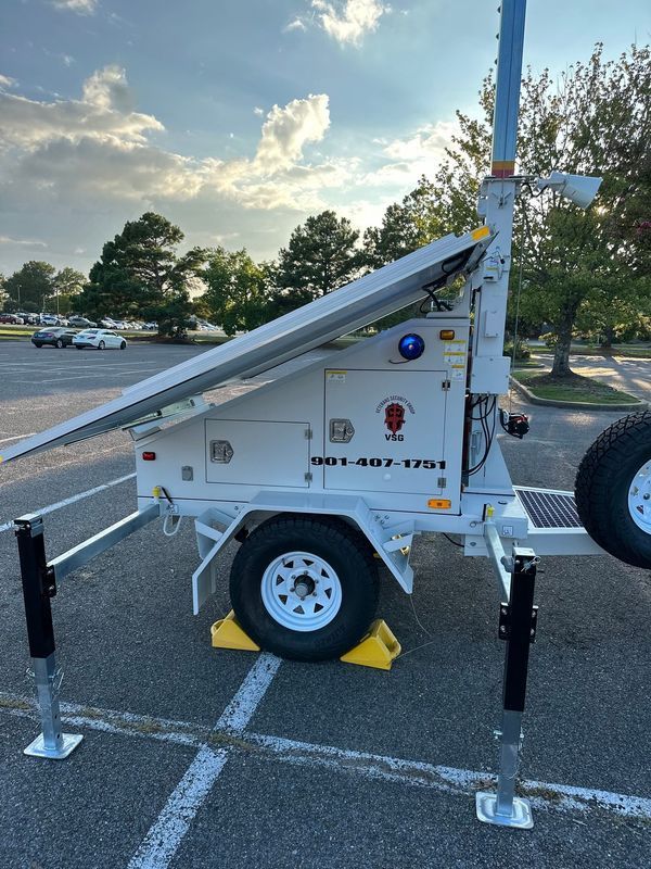 White solar-powered trailer with raised solar panel and extendable legs, parked on asphalt, under a cloudy sky.