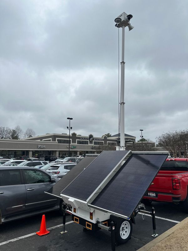 A security camera trailer with solar panels in a parking lot on a cloudy day. Cameras are mounted on a telescoping pole.