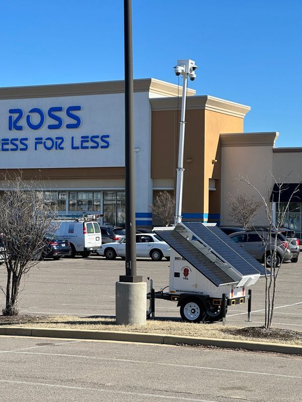 Mobile security unit with solar panels and cameras in a parking lot in front of a Ross store. Clear blue sky.