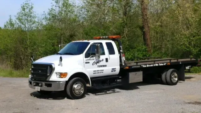 White flatbed tow truck parked on a gravel road beside trees, side view with orange beacon lights.