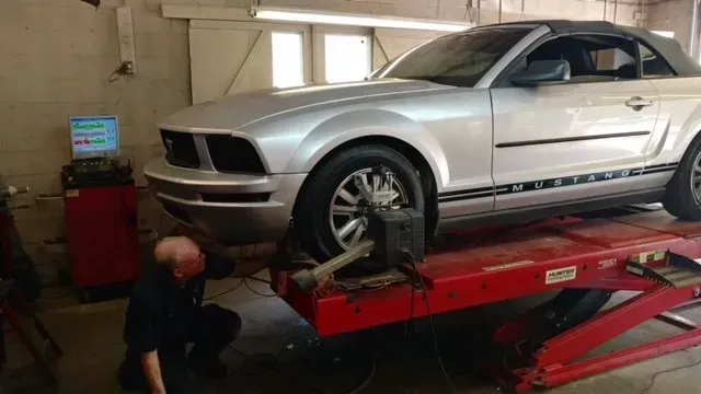 Silver Mustang on a red car lift in an auto shop, with a mechanic working below.