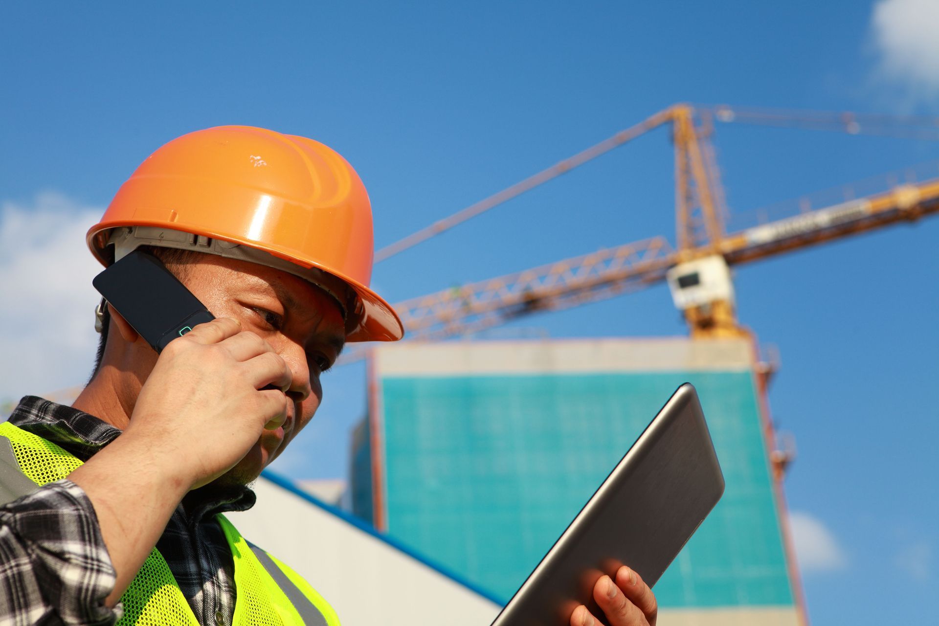 Construction worker wearing a hard hat, talking on a phone, holding a tablet, with a building and crane in the background.