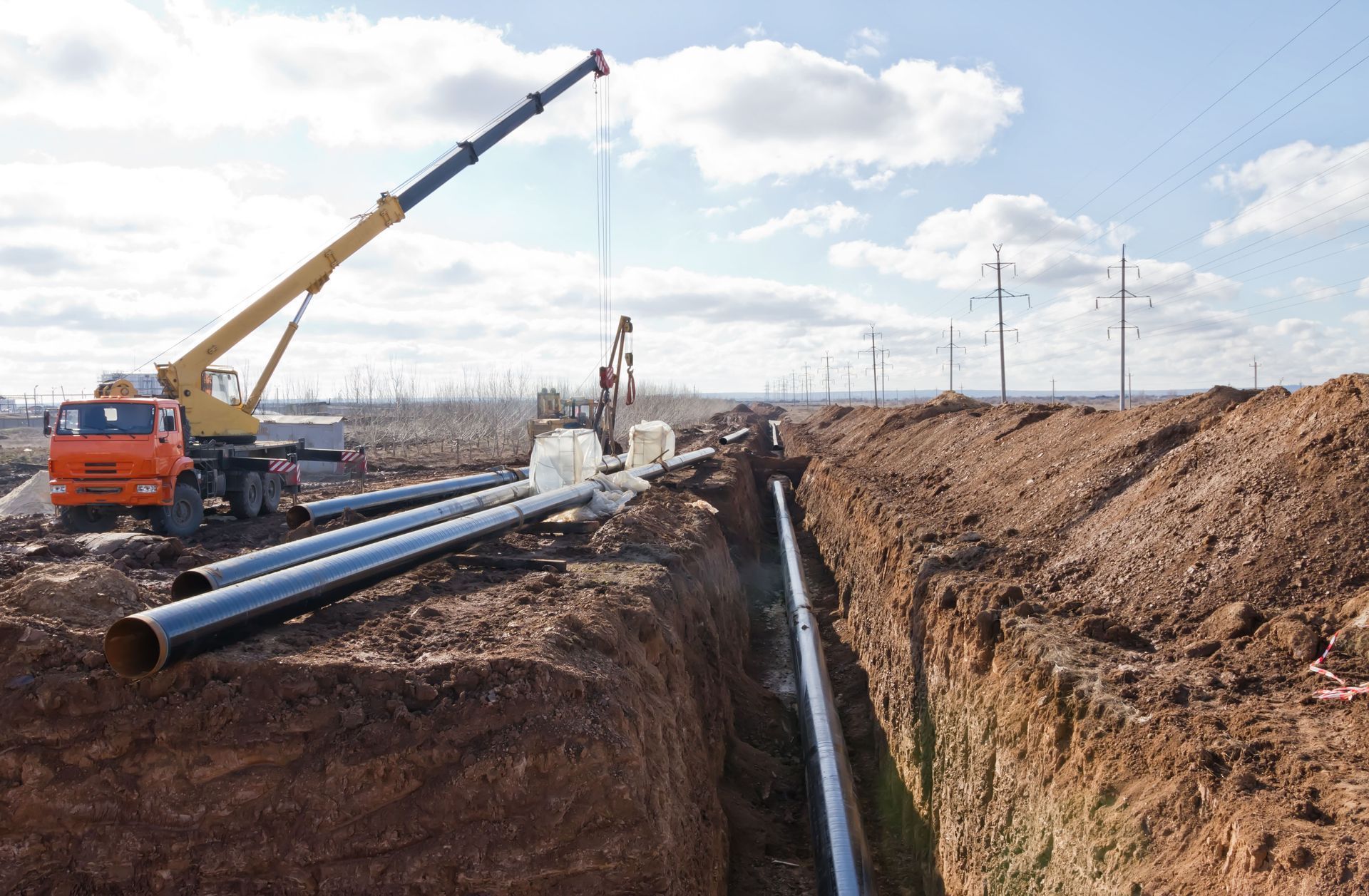 Crane lowering a pipe section into a trench for pipeline construction outdoors under a partly cloudy sky.