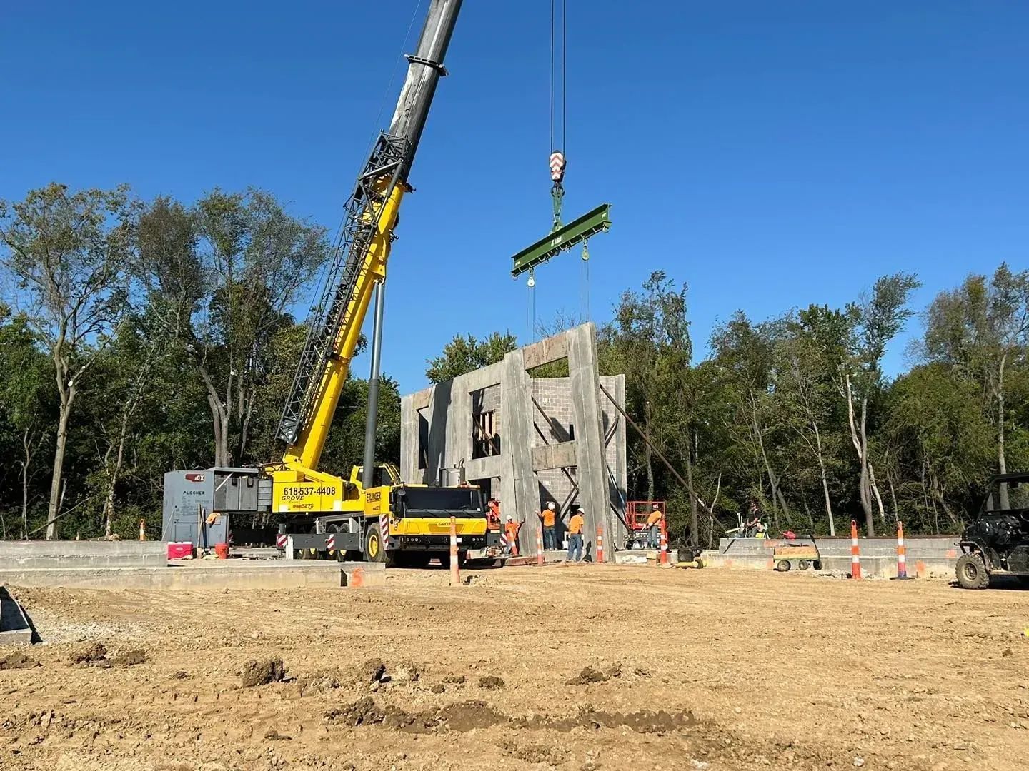 Construction site with a crane lifting a concrete wall section. Workers in orange vests. Clear blue sky.