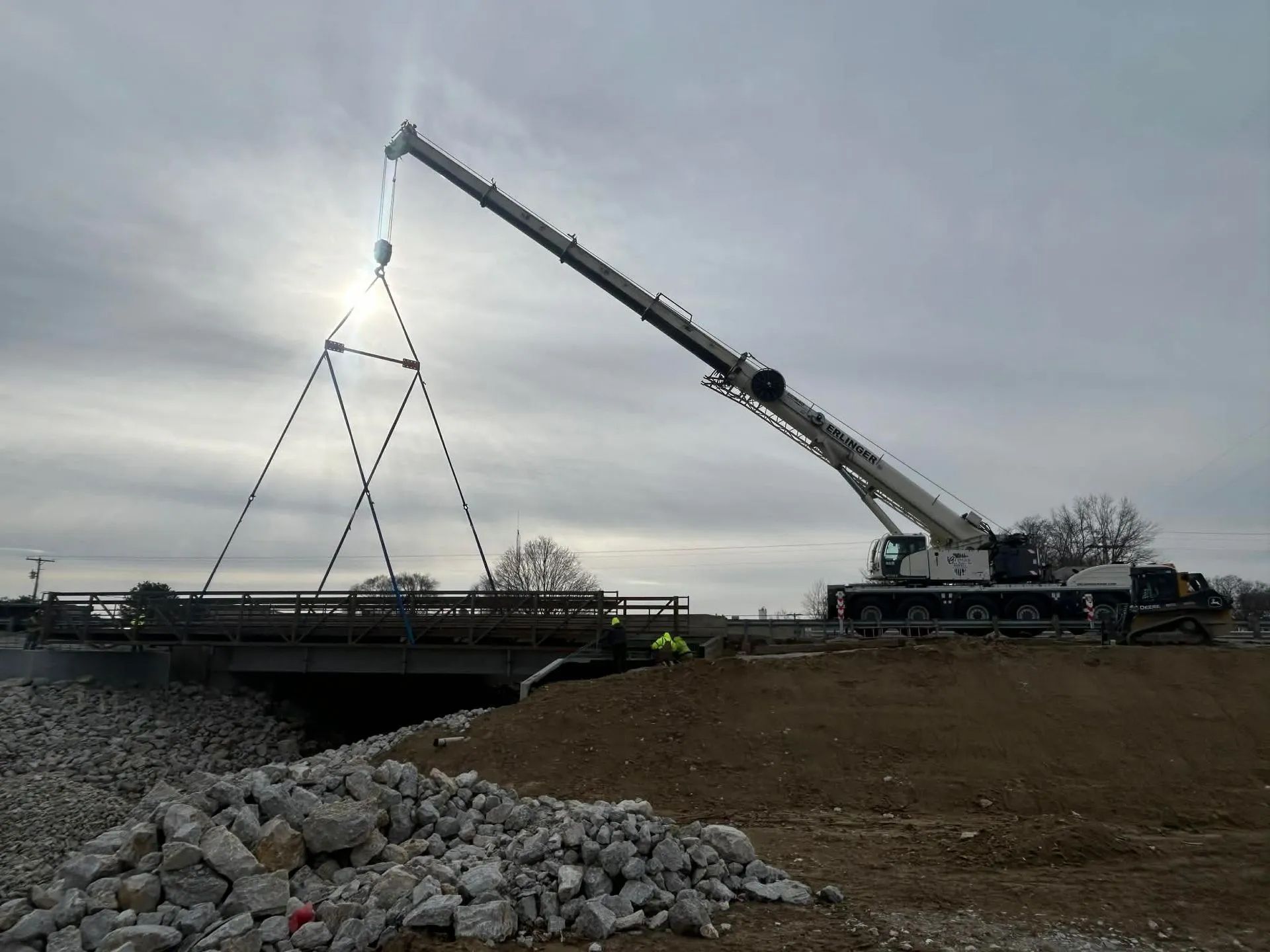 Crane lifting a bridge section over a waterway; construction site with dirt, rocks, and overcast sky.