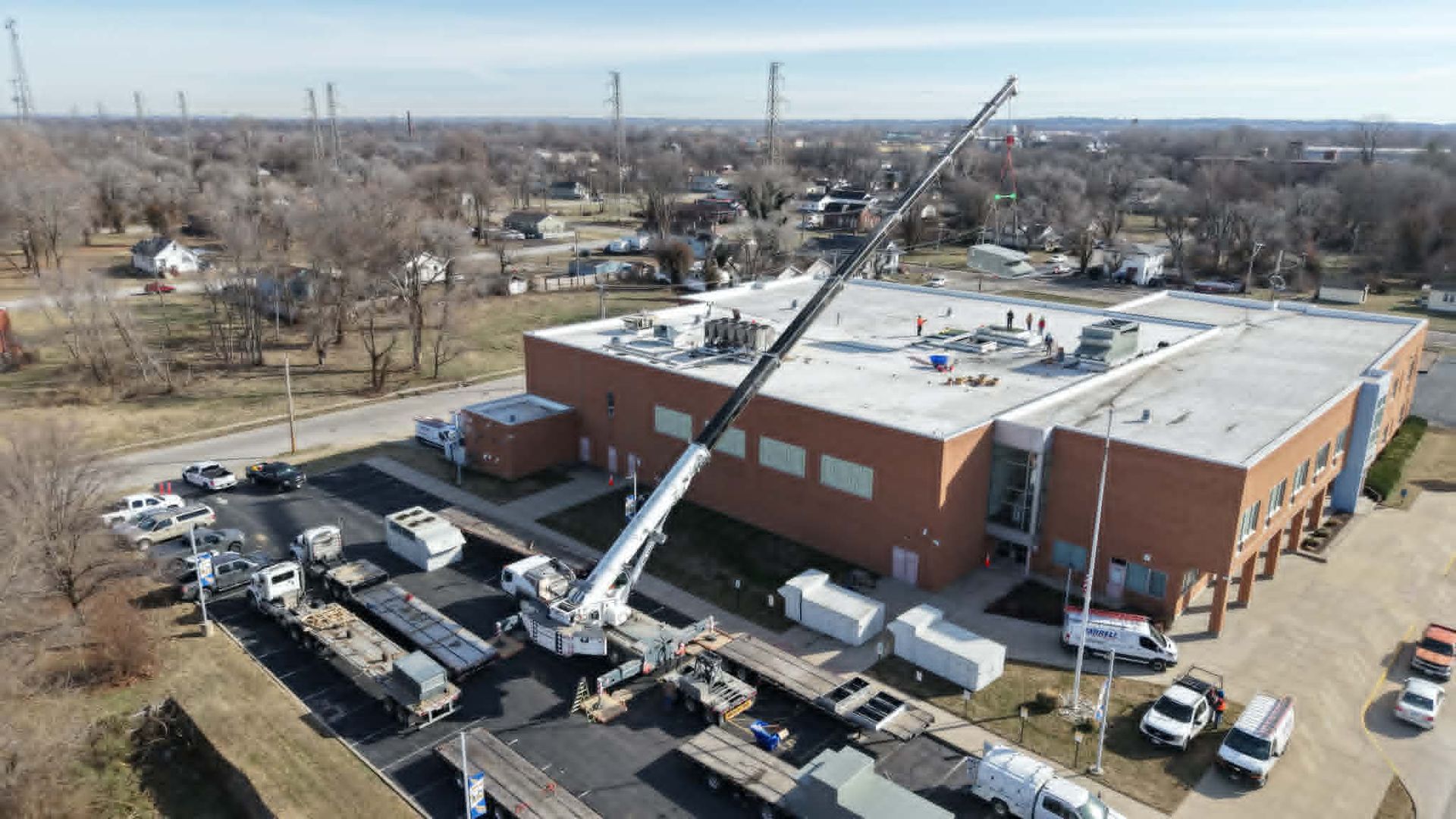 A crane lifts equipment onto a red brick building roof in an urban setting. Numerous trucks are parked nearby.