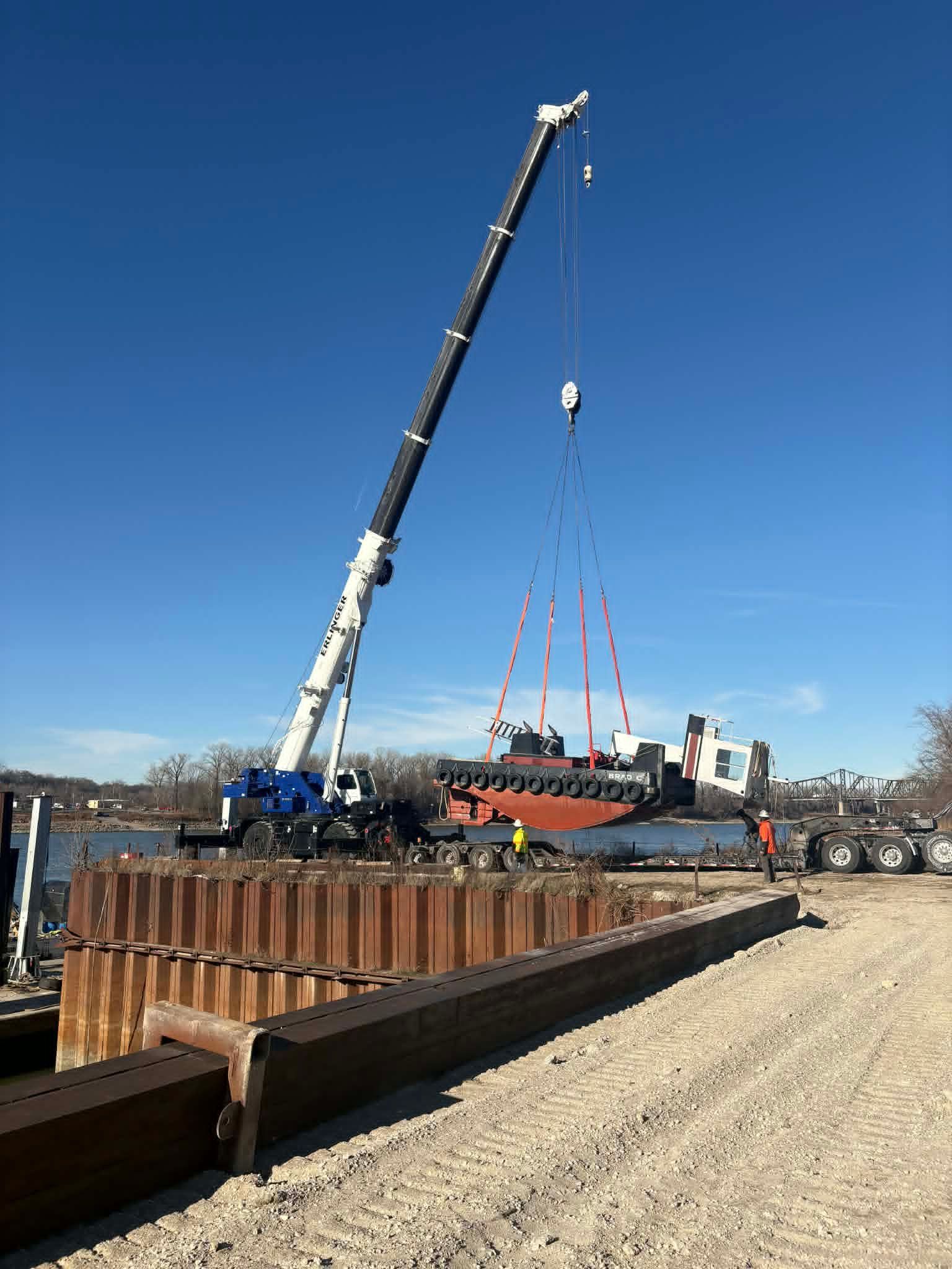 A crane lifts a large industrial component over a construction site near water on a sunny day.