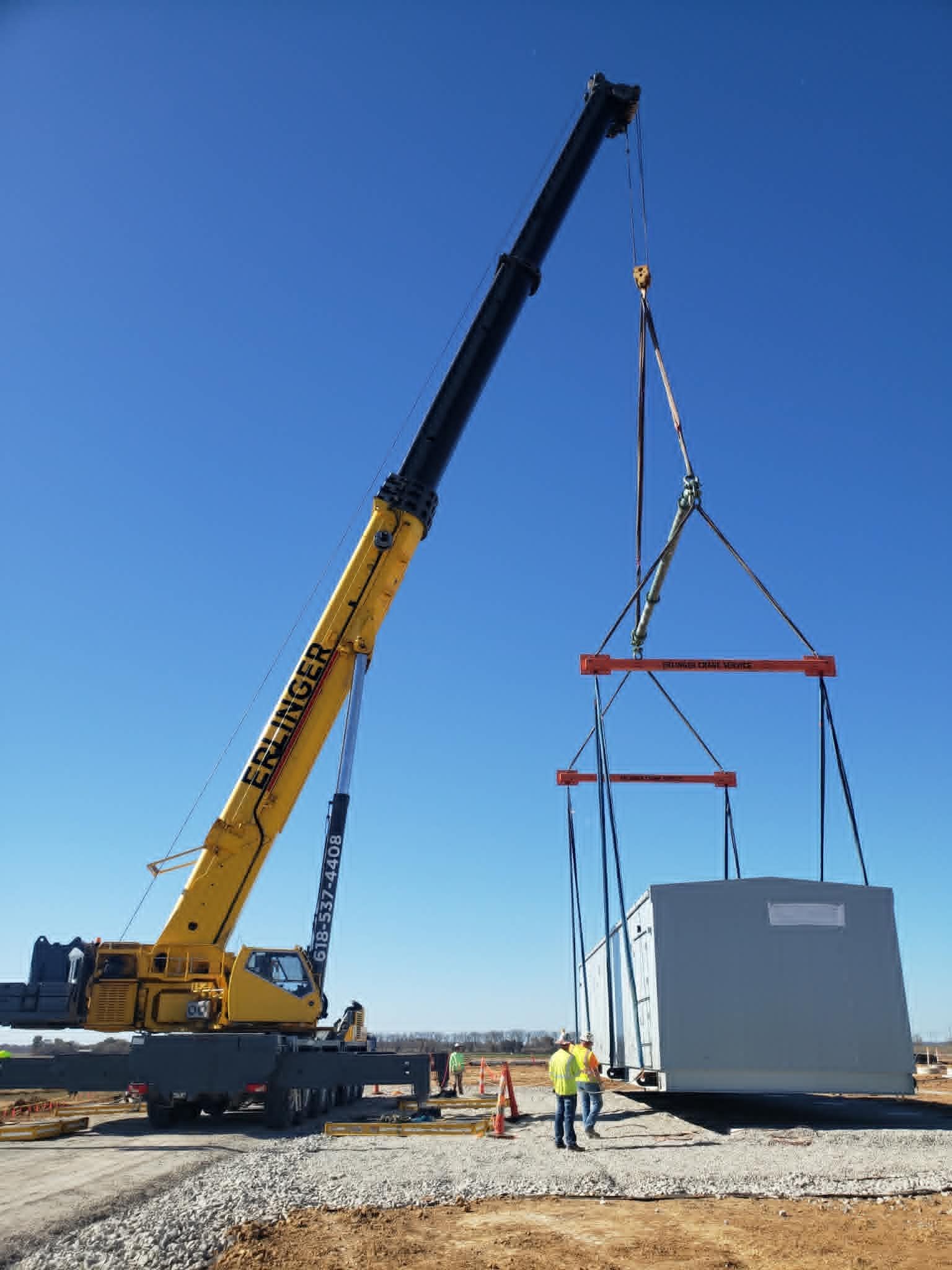 A large yellow crane lifting a gray rectangular structure, two workers watching in a sunny outdoor setting.