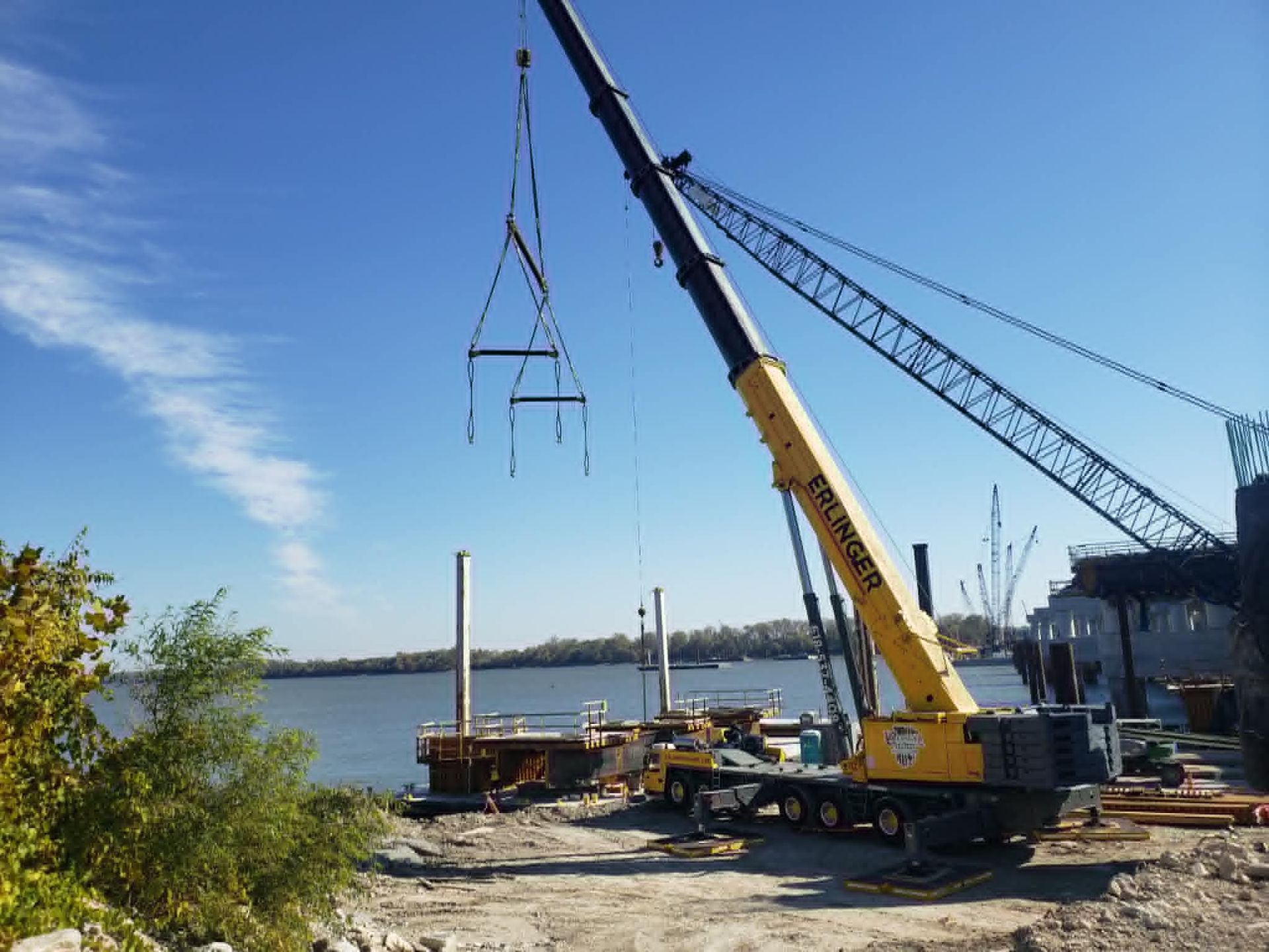 A large yellow crane lifting metal framework near a river. Construction site on a sunny day.